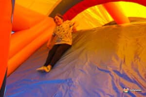 A child in a patterned shirt slides down a colorful inflatable surface inside a bouncy castle, surrounded by bright hues of orange and yellow.