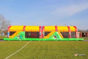 A colourful inflatable ultimate obstacle course with a slide, set on a grassy field under a clear blue sky, perfect for outdoor fun.