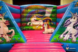 Colorful inflatable bouncy castle featuring playful unicorns, vibrant rainbows, and butterflies on a bright, grassy landscape.