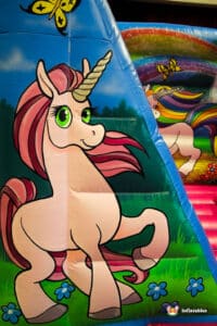 Colourful cartoon unicorn with pink mane and big green eyes surrounded by flowers and butterflies on an inflatable slide backdrop.