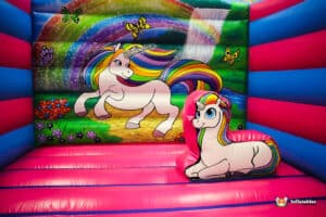 A colourful bouncy castle features a vibrant unicorn mural and a plush unicorn cushion, set against a whimsical landscape.