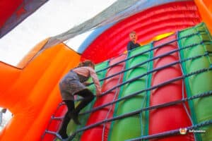 A child climbing a colorful inflatable wall inside a bouncy castle, with bright orange, red, and green hues visible.