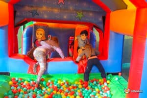 Two children play in a colorful inflatable bounce house filled with vibrant plastic balls, climbing toward the entrance.