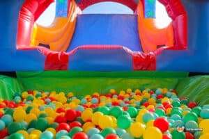 A colorful inflatable bounce house with a slide, filled with a variety of vibrant plastic balls in a green pit below.