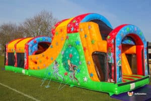 A colourful inflatable wedge slide ball pool fun run with a slide, featuring stars and cartoon characters, set on a grass field under a clear blue sky.