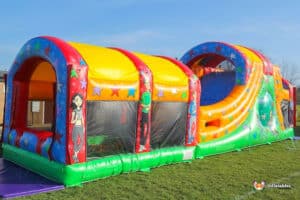 A colourful inflatable wedge slide ball pool fun run with a slide, decorated with stars and cartoon characters, set on green grass under a clear blue sky.