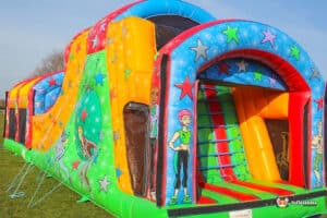 A colourful inflatable wedge slide ball pool fun run with bright stars and cartoon figures, featuring slides and climbing areas, set on lush grass.