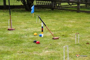 Croquet setup on green lawn, featuring a mallet, colorful balls, and wickets, with a blue flag marking the playing area.