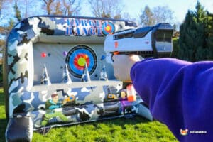 A person aims a toy blaster at an inflatable target range featuring colorful targets and camouflage design set on grass.