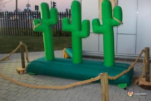 An inflatable green cactus structure surrounded by rope and wood, set against a twilight sky and grass backdrop.