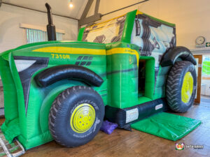 Angled view of a large, custom-made, inflatable John Deere 7310R tractor bouncy castle set up indoors on a wooden floor, featuring realistic green and yellow coloring and large inflatable tires.