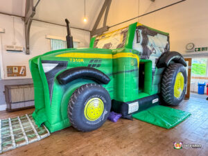 Frontal view of the inflatable tractor bouncy castle in a large hall. The interior bouncing area is visible from the side opening near the large yellow-rimmed back tire.