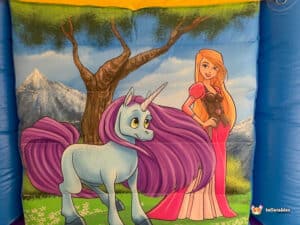 A princess stands beside a unicorn bouncy castle, smiling with her horse in a vibrant, playful setting
