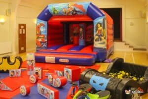A colorful inflatable bouncy castle with superhero motifs, accompanied by an assortment of toys and playful accessories.