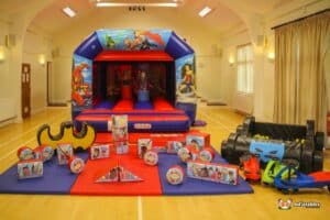 A large inflatable bouncy castle decorated with superhero themes and surrounded by colorful toys and play items.