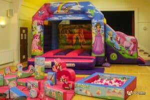 A colorful inflatable bounce house with various princesses and unicorns, surrounded by playful foam blocks and a ball pit in a spacious room.