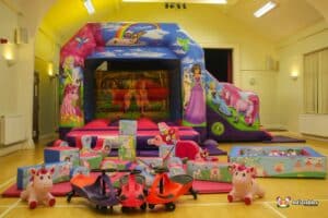 A colorful inflatable castle with princesses and unicorns, surrounded by play mats and toy vehicles in a bright indoor space.