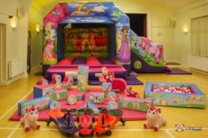 A colorful indoor playground featuring a large inflatable bouncy castle, soft play blocks, ride-on toys, and a ball pit.