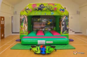 A colorful bouncy castle featuring jungle animals, with pink and green inflatable surfaces, and toy ride-on vehicles in front.