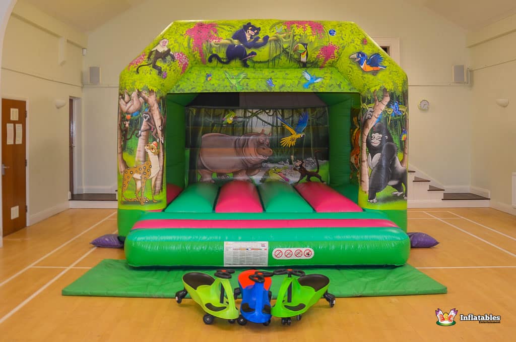 A colorful bouncy castle featuring jungle animals, with pink and green inflatable surfaces, and toy ride-on vehicles in front.