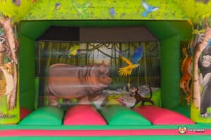 Colourful inflatable bouncy castle featuring a cartoon hippo surrounded by jungle animals like monkeys and parrots in a vibrant setting.