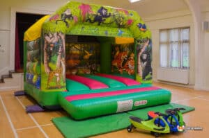 A colourful inflatable bouncy castle featuring jungle animals, with vibrant pink and green surfaces, and children's ride-on toys beside it.