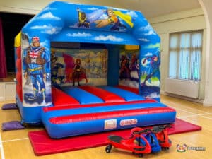 A red and blue bouncy castle with a hero theme, featuring plasma cars for kids to play on and enjoy.