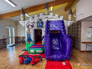 A colorful inflatable castle with slides, featuring playful characters, set in a spacious indoor area with chairs and a safety mat.