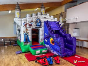 A colorful inflatable castle with a dragon slide and whimsical characters, set indoors with chairs in the background.