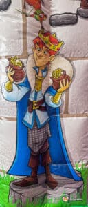 A cheerful cartoon king with red hair and a crown holds two pots of gold, wearing a blue coat and standing on a grassy stone pedestal.