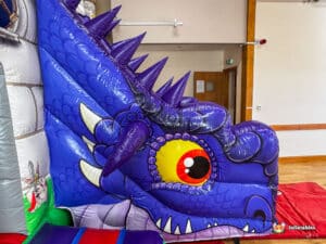 A colorful inflatable dragon's head with a striking yellow eye and purple scales, featured in a vibrant indoor bounce area.