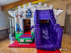 A colorful inflatable bouncy castle with a slide, featuring playful medieval-themed graphics, set up indoors with wooden beams overhead.