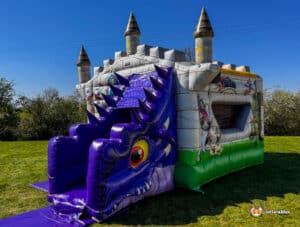 Inflatable dragon-themed bounce house with a colorful castle design and slide, set in a grassy area under a blue sky.