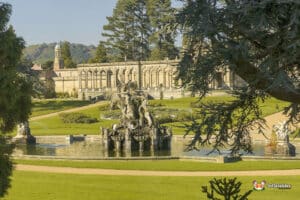 A grand garden view featuring a classical fountain, lush green lawns, and an ornate building in the background under clear blue skies.
