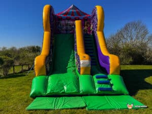 A large inflatable circus slide with vibrant colours, featuring a striped canopy, green slide, and blue stairs, set against a clear blue sky.