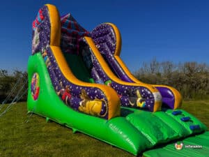 A colourful inflatable circus slide with two parallel slides, featuring cartoon characters and a bright green base, set against a clear blue sky.