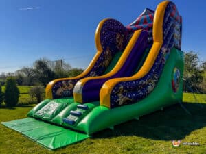 A colourful inflatable circus slide with two lanes, set in a sunny outdoor field, featuring cartoon illustrations and green grass below.