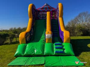 A colourful inflatable circus slide featuring a steep slide and stairs, set on a green lawn under a clear blue sky. Fun outdoor attraction for kids.