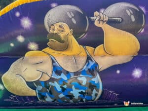 A cartoon muscular figure in a blue camouflage tank top lifts a heavy barbell, set against a starry, colorful background.