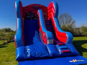 A colourful inflatable super hero slide featuring superhero designs, with two side slides and steps, set on a bright, sunny day outdoors.
