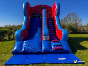 Bright red and blue inflatable super hero slide with superhero characters, featuring two steep slides and a staircase, set in a grassy area.