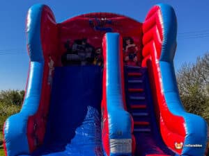 A vibrant inflatable slide featuring superhero designs, including Spider-Man, Batman, and Wonder Woman, set against a clear blue sky.