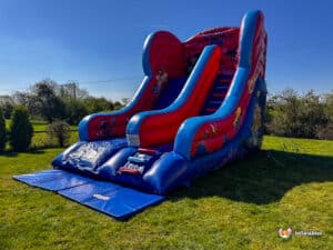 A vibrant inflatable super hero slide featuring cartoon characters, set on green grass under a clear blue sky. Perfect for outdoor fun!