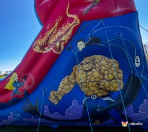 A colorful inflatable slide featuring comic book characters: a female superhero, a fiery figure, and The Thing, all set against a blue background.