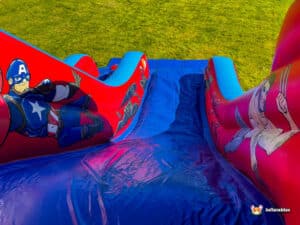 A brightly colored inflatable slide featuring superhero designs, including Captain America and Wolverine, on a grassy outdoor setting.