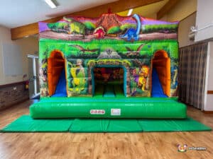 A colorful dinosaur-themed inflatable double slide cabin with vibrant characters and a slide, set in a spacious indoor area.