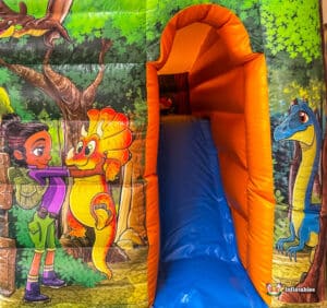 Colorful inflatable with a girl and dinosaur illustration, featuring a blue slide surrounded by vibrant forest scenery.