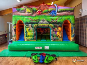 A colorful dinosaur-themed dinosaur double slide cabin with vibrant illustrations, set indoors on a green inflatable base, with ride-on toys nearby.