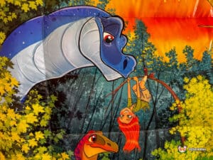 A vibrant cartoon scene featuring a blue dinosaur, a hanging girl with red hair, and a playful dinosaur in a lush forest setting.