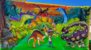 A vibrant dinosaur-themed backdrop featuring various cartoon dinosaurs, lush greenery, and a child interacting with them.